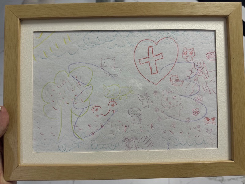 Coloured line drawing filling the page: cats, small figures, a tree, flowers, a large red heart with a cross inside, and rows of waves along the top and bottom.